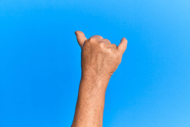 Hand of senior hispanic man over blue isolated background gesturing hawaiian shaka greeting gesture, telephone and communication symbol  - Photo, Image