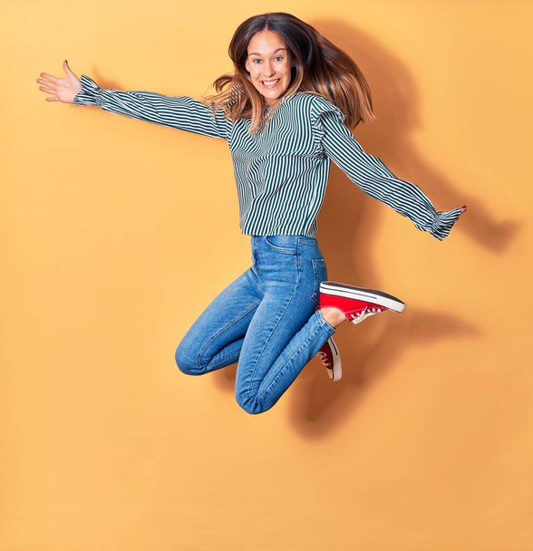 Young beautiful caucasian woman wearing casual clothes smiling happy. Jumping with smile on face and arms opened over isolated yellow background - Фото, зображення