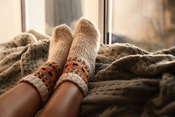 Woman in knitted socks relaxing on plaid near window at home, closeup - Foto, imagen