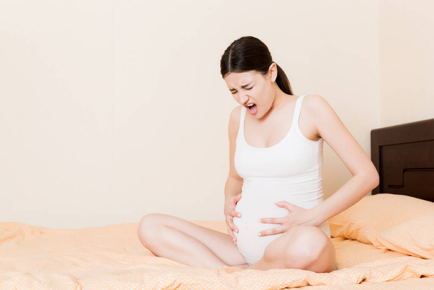 Беременная женщина с массажем нижнего живота сидит на диване в помещении. Концепция проблем с беременностью. Материнство. - Фото, изображение