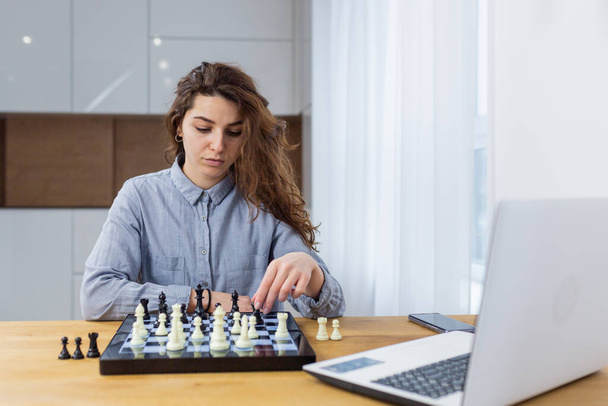 Beautiful girl sitting at home and playing chess online with a laptop - 写真・画像