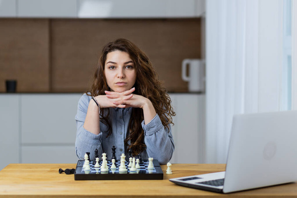 Beautiful girl sitting at home and playing chess online with a laptop - Foto, Imagem