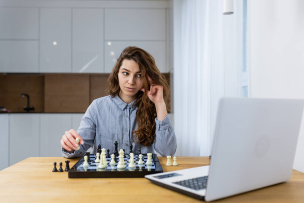 Beautiful girl sitting at home and playing chess online with a laptop - Fotó, kép