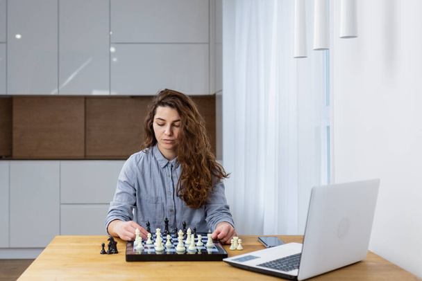 Beautiful girl sitting at home and playing chess online with a laptop - Photo, Image