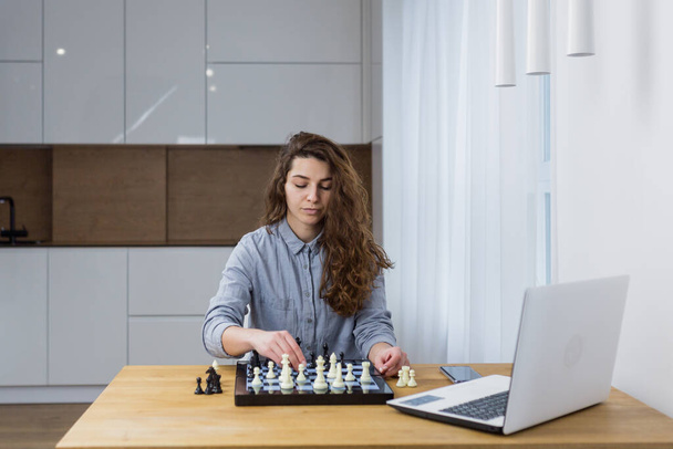 Beautiful girl sitting at home and playing chess online with a laptop - Fotoğraf, Görsel