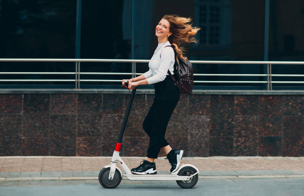Fiatal nő mosollyal lovagolni elektromos robogó az utcán - Fotó, kép