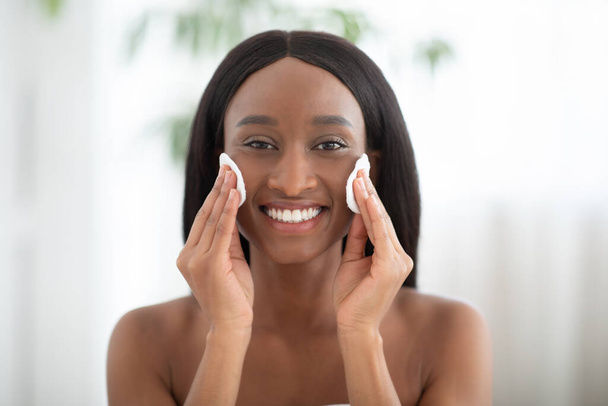 Souriante jeune femme afro-américaine qui pratique quotidiennement des soins de la peau le matin, nettoie la peau du visage avec une lotion naturelle - Photo, image