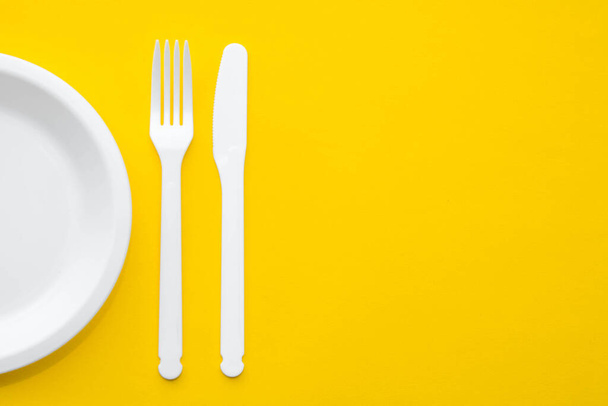 Plastic white fork, knife and plate on yellow background. Cooking utensil. Top view. Minimalist Style. Copy, empty space for text. - Photo, Image
