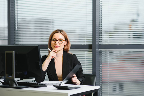 Pretty, nice, cute, perfect woman sitting at her desk on leather chair in work station, wearing glasses, formalwear, having laptop and notebook on the table - Фото, изображение