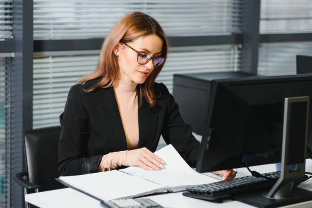 Pretty, nice, cute, perfect woman sitting at her desk on leather chair in work station, wearing glasses, formalwear, having laptop and notebook on the table - Photo, image