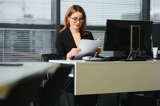 Pretty, nice, cute, perfect woman sitting at her desk on leather chair in work station, wearing glasses, formalwear, having laptop and notebook on the table - Photo, image