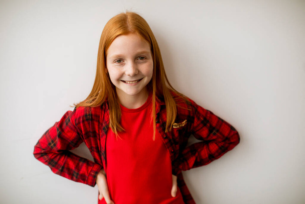 Linda niña de pelo rojo de pie junto a la pared blanca - Foto, Imagen