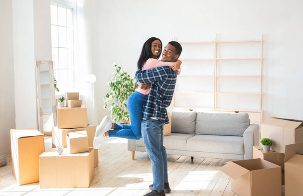 Happy moving day. Loving black man embracing and lifting his pretty girlfriend up in their new home, panorama - Fotografie, Obrázek