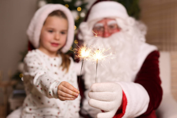 Santa Claus and little girl in room decorated for Christmas, focus on burning sparklers - Fotó, kép