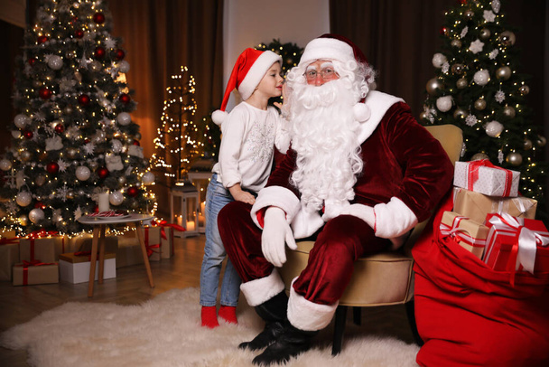 Little girl whispering in Santa Claus' ear near Christmas tree indoors - Photo, Image