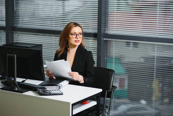 Young businesswoman sitting at workplace and reading paper in office - Foto, Bild