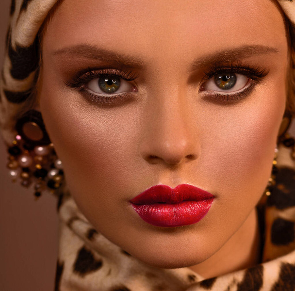 Female portrait. Face close-up. Hat and scarf in a leopard pattern. Large earrings in the ears. Red lipstick. - 写真・画像