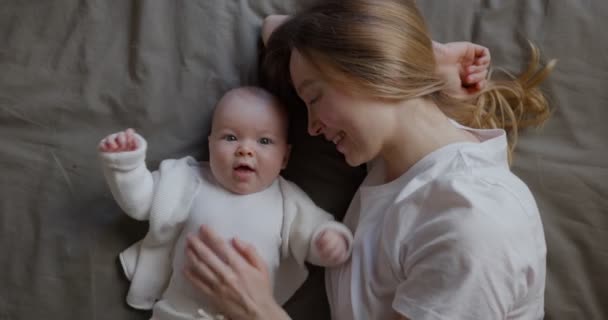 Mom with her baby lies and looks at the camera from the top. Beautiful young woman with her baby. Mother and daughter. Happy family. - Footage, Video