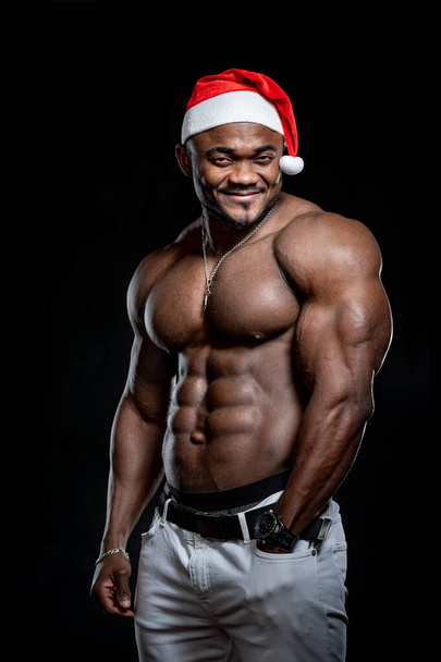 Smiling bodybuilder with strong body poses to the camera with naked torso. Athlete wears santa hat. Black background. - Fotoğraf, Görsel