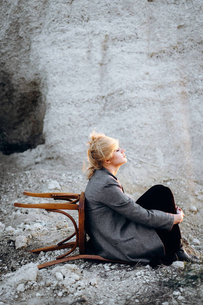 Chica pelirroja sentada en una silla vieja en un cañón de arena al atardecer. Retrato psicológico de la soledad. Hipster viejo concepto de moda - Foto, Imagen