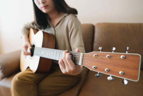 Beautiful asian woman playing acoustic guitar while sitting on a couch at home. Hobbies and leisure activities concept. Selective focus. - Photo, image