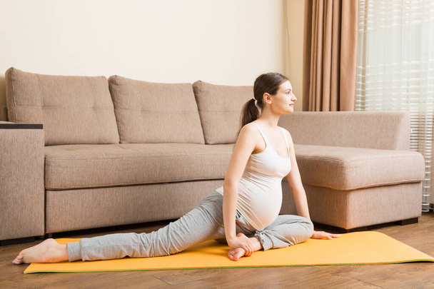 gyönyörű fiatal terhes nő ül kanapé mellett jóga pózban otthon. Terhesség Jóga és Fitness koncepció a koronavírus idején. - Fotó, kép