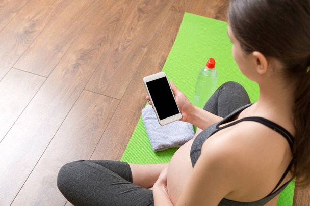 Schwangere hält Smartphone in der Hand, während sie zur Zeit des Coronavirus zu Hause auf der Yogamatte sitzt. - Foto, Bild