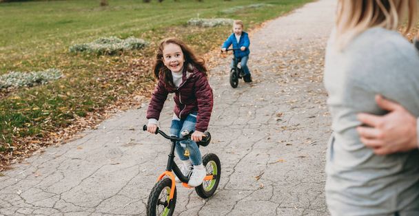 Valkoihoinen tyttö ja hänen veljensä ratsastus pyörä puistossa, kun katsot heidän vanhempansa syleilevät - Valokuva, kuva