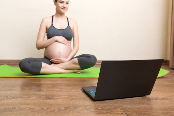 Молодая счастливая здоровая фитнес yoga красивейшая беременная женщина делает тренировку yoga bakasana на циновке yoga напротив ноутбука с online мастер-классом. концепция образа жизни в коронавирусное время. - Фото, изображение