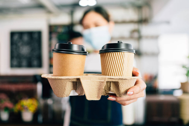 喫茶店では、フェイスマスクをした女性バリスタが紙コップでコーヒーを出している。コロナウイルスのパンデミックの間の小さな緑のビジネス。クライアントの所有者と従業員サービス. - 写真・画像
