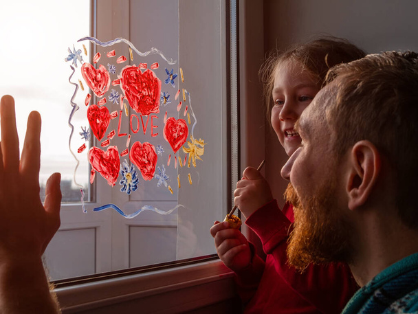 Child with father painting Love sign, playing indoors quarantine family leisure. Little girl holds paintbrush in hand draws red heart on window glass. Valentine's day. Stay home art concept New normal - Photo, Image