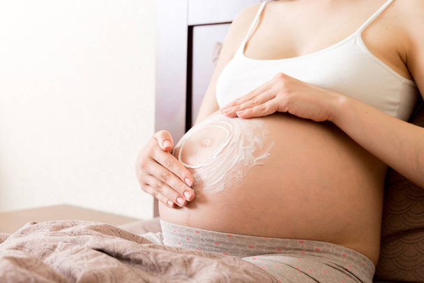 a pregnant girl sits at home on the bed and smears an anti-stretch mark cream on her stomach. Pregnancy, motherhood, preparation and expectation concept. - Photo, Image