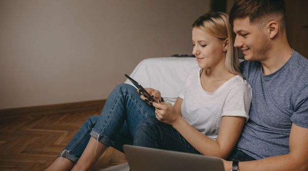 Preciosa pareja caucásica sentada en el suelo y usando un ordenador portátil y una tableta - Foto, Imagen