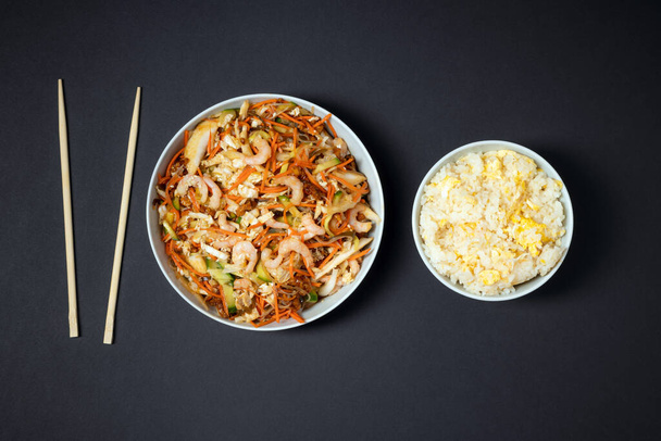 lebendiger Teller mit Garnelensalat, Schüssel Reis mit Ei und Essstäbchen auf schwarzem Hintergrund. Essen mitnehmen. - Foto, Bild