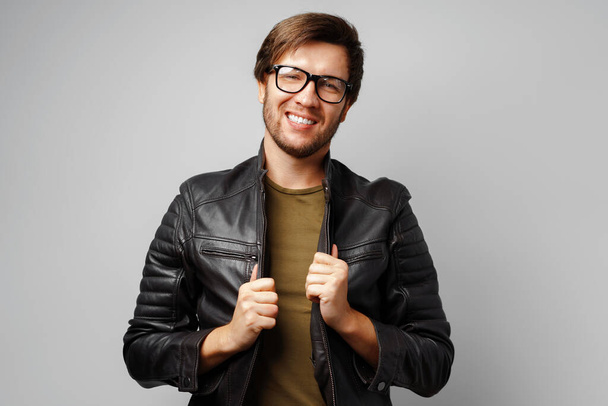 Portrait of a young man in glasses wearing black leather jacket on grey background - Photo, Image