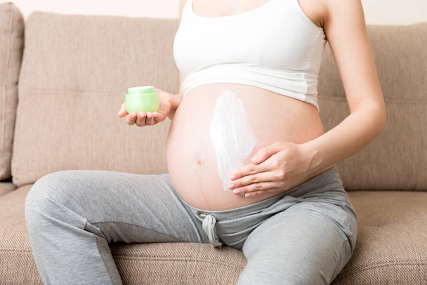 una chica embarazada se sienta en casa en la cama y frota sonrisa una crema anti-estrías en su estómago. Concepto de embarazo, maternidad, preparación y expectativa. - Foto, Imagen