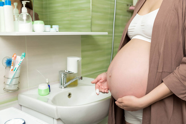 Young pregnant woman applying anti-stretch marks cream to the abdomen in the morning in the bathroom. Pregnancy, maternity and bodycare concept. - Zdjęcie, obraz