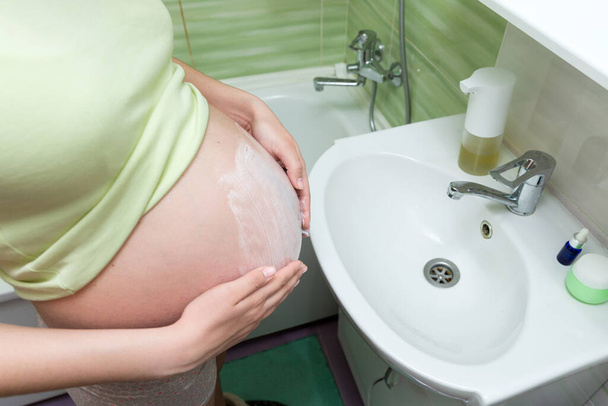 Junge Schwangere cremen morgens im Badezimmer den Bauch mit Dehnungsstreifen ein. Schwangerschaft, Mutterschaft und Körperpflege-Konzept. - Foto, Bild