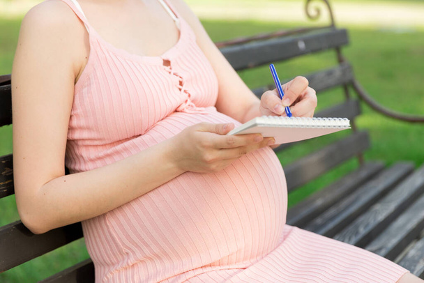 těhotná žena sedí na lavičce v parku a píše seznam úkolů do nemocnice. - Fotografie, Obrázek