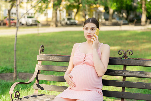 donna incinta siede su una panchina del parco e parla al telefono. - Foto, immagini