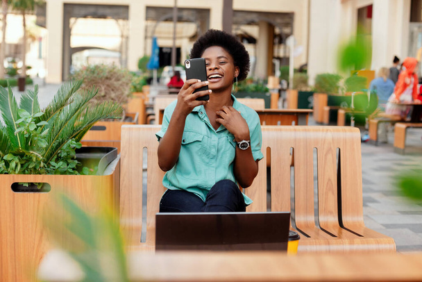 Una giovane ragazza africana con gli occhiali scatta una foto di selfie sul suo smartphone, posando davanti alla fotocamera. Una ragazza si fa un selfie durante una pausa mentre lavora su un computer portatile. - Foto, immagini