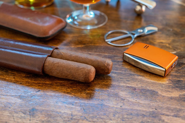 Сигара и аксессуары на деревянном столе, вид крупным планом. Кубинские сигары и бренди. Табак и алкоголь, курение и питье роскошный образ жизни - Фото, изображение