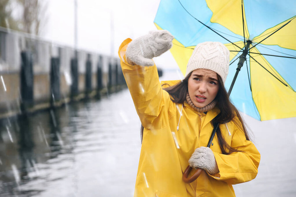 Woman in yellow raincoat with umbrella caught in gust of wind near river - Фото, изображение