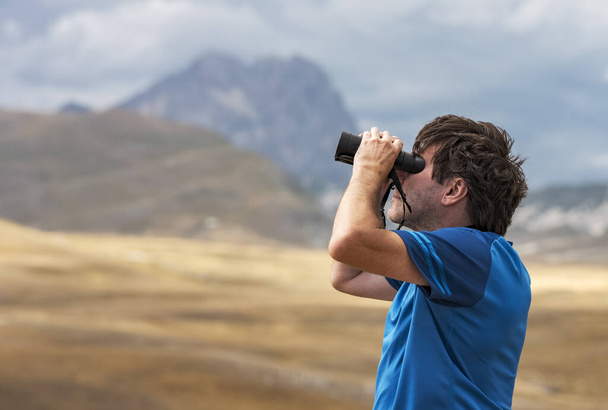 Portrait of a man standing on a mountain hill and looking into the binoculars in the distance, Campo Imperatore, Gran Sasso National Park, Abruzzo region, Italy. Travel concept. Concept of person looking to the future.  - Photo, Image