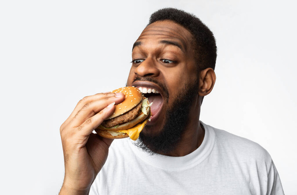 Portrait d'un homme noir affamé mangeant un hamburger sur fond blanc - Photo, image