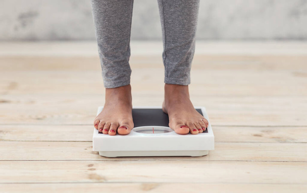 Крупный план молодой афроамериканской женщины, стоящей на весах в помещении, проверяющей свой вес, обрезанной вид на ноги - Фото, изображение