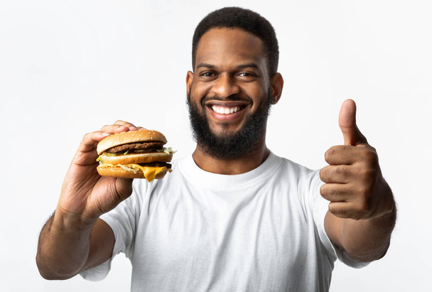 Black Man Holding Burger And Gesturing Thumbs-Up Over Yellow Background - Foto, Imagen