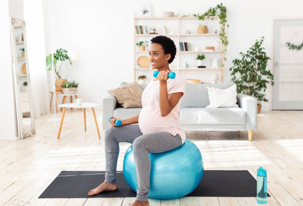 Pregnant black woman sitting on fitness ball, working out with dumbbells in her home gym - Foto, Imagem