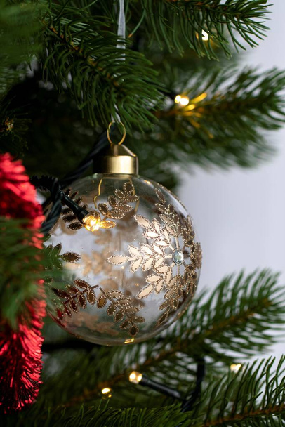 Portret szklanej piłki bożonarodzeniowej ozdobionej złotem i diamentami zwisającymi z choinki wraz z innymi ozdobami świątecznymi, takimi jak lampki świąteczne. - Zdjęcie, obraz