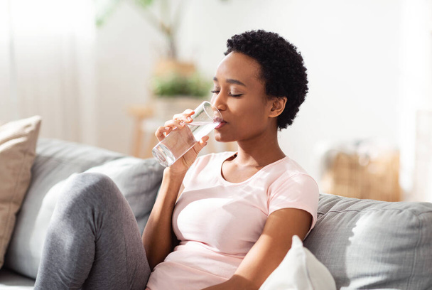 Hydratace pro dobrý zdravotní koncept. Hezká mladá černoška se sklenicí křišťálově čisté vody na gauči doma - Fotografie, Obrázek